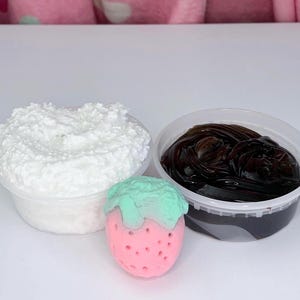 May include: Two clear plastic containers filled with textured substances. One container holds a white, grainy, foam-like material, while the other contains a dark brown, glossy, translucent gel. A small pink strawberry-shaped object with a mint green top sits between them.