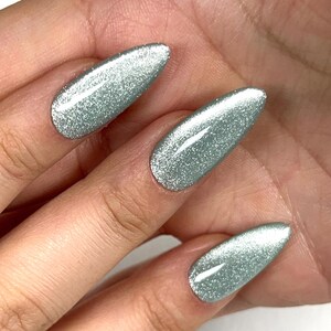 Pode incluir: Close-up de três unhas com um verniz verde claro e brilhante. As unhas são ovais e refletem a luz, criando um efeito brilhante. O fundo é uma visão desfocada da pele.