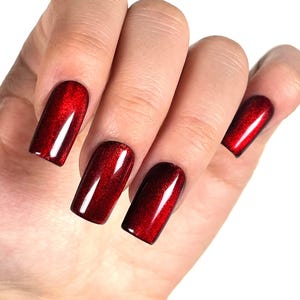 Peut inclure: Gros plan d'une main avec des ongles vernis. Les ongles sont d'un rouge profond avec une finition brillante. Les ongles sont de forme rectangulaire et reflètent la lumière, créant un effet chatoyant. Le fond est blanc.