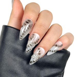 Puede incluir: Una mano que muestra uñas artificiales largas y puntiagudas. Las uñas presentan una base blanca con patrones grises ahumados y detalles de cadena plateada. Una uña tiene un adorno floral plateado.