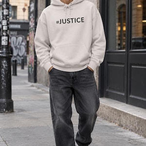 Op de afbeelding: Een lichtbeige hoodie met de tekst #JUSTICE in zwarte letters op de borst, gecombineerd met een ruimvallende antracietgrijze spijkerbroek en witte sneakers. De hoodie is voorzien van een kangoeroezak en trekkoorden bij de capuchon.