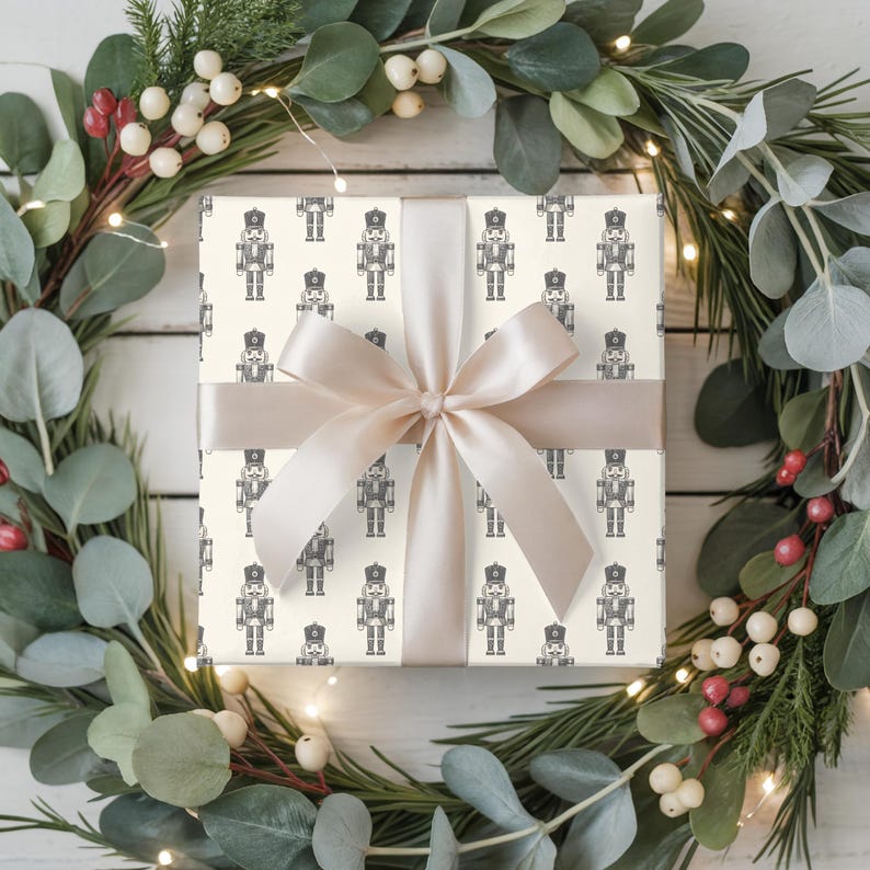A top-down view of a wreath made of evergreen branches, eucalyptus, and red and white hypericum berries sit on a white painted wood plank floor. White fairy lights are strung around the wreath. A wrapped gift sits in the middle of the wreath. The gift is wrapped with a repeating pattern of traditional nutcrackers. The gift wrap is cream and the nutcracker pattern is charcoal gray. The gift is wrapped with a white satin ribbon and is tied in a bow at the center.