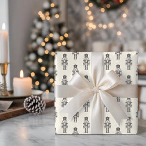 A wrapped package sits on a marble table. The wrapping paper is cream with a repeating pattern of traditional nutcrackers in charcoal gray and tied with a cream satin ribbon and bow. A wooden board with a lit white taper in a bronze candlestick, pinecones, and lit white pillar candle sits the left. The room has light gray walls and a gray wallpaper. A flocked Christmas tree with white lights sits in the corner, and a wreath are partially shown on the wallpapered wall.