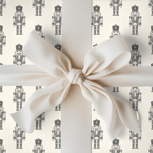 A tight close-up view of a wrapped gift. The gift fills the entire frame of the image. The wrapping paper is a repeating pattern of an etched-style traditional nutcrackers. The wrapping paper color is cream and the nutcracker pattern is a charcoal gray. The gift is wrapped with a white ribbon and tied in a bow at the center.