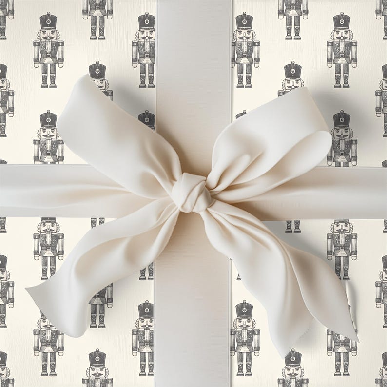 A tight close-up view of a wrapped gift. The gift fills the entire frame of the image. The wrapping paper is a repeating pattern of an etched-style traditional nutcrackers. The wrapping paper color is cream and the nutcracker pattern is a charcoal gray. The gift is wrapped with a white ribbon and tied in a bow at the center.