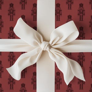 A tight close-up view of a wrapped gift. The gift fills the entire frame of the image. The wrapping paper is a repeating pattern of etched-style traditional nutcrackers. The wrapping paper color is burgundy and the nutcracker pattern is a darker shade of burgundy. The gift is wrapped with a white ribbon and tied in a bow at the center.