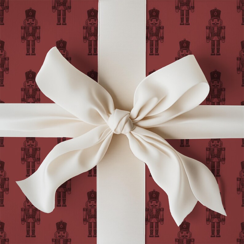 A tight close-up view of a wrapped gift. The gift fills the entire frame of the image. The wrapping paper is a repeating pattern of etched-style traditional nutcrackers. The wrapping paper color is burgundy and the nutcracker pattern is a darker shade of burgundy. The gift is wrapped with a white ribbon and tied in a bow at the center.