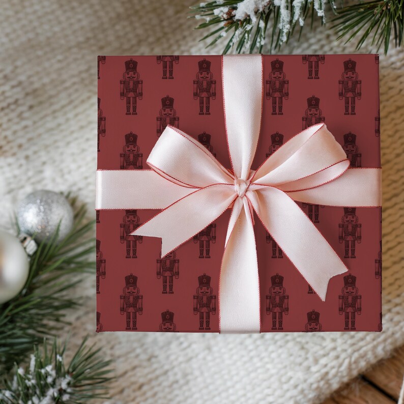 A top-down view of a gift sitting on a cream knit blanket lying on a medium toned wood floor. Flocked greenery and satin finish white and silver Christmas ornaments surround the gift. The present is wrapped in a repeating pattern of an etched-style nutcracker. The wrapping paper is burgundy and the nutcracker pattern is a darker shade of red. The gift is wrapped in a light pink satin ribbon with a darker pink edge. The gift is tied with a bow at the center.