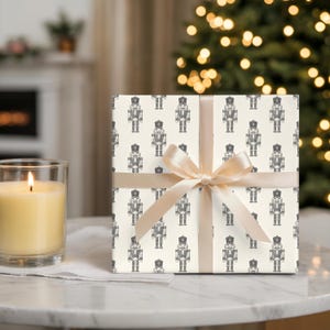 A wrapped gift sits on a marble tabletop with a cream-colored lit candle in a glass jar to the left. The wrapping paper features a repeating pattern of etched style traditional nutcrackers. The wrapping paper is a cream, and the nutcracker pattern is in charcoal gray. The gift is wrapped with a thin, cream satin ribbon. Out of focus in the background, a white fireplace with greenery on the mantel.