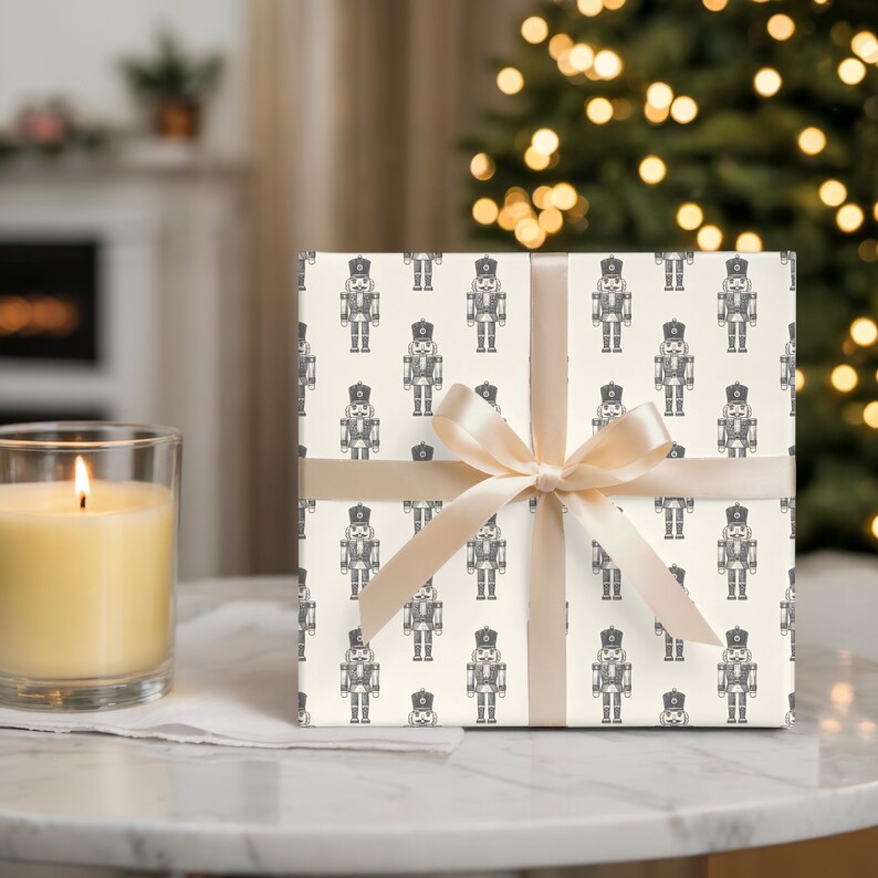 A wrapped gift sits on a marble tabletop with a cream-colored lit candle in a glass jar to the left. The wrapping paper features a repeating pattern of etched style traditional nutcrackers. The wrapping paper is a cream, and the nutcracker pattern is in charcoal gray. The gift is wrapped with a thin, cream satin ribbon. Out of focus in the background, a white fireplace with greenery on the mantel.