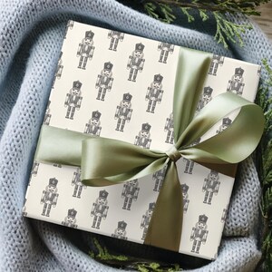 A top-down view of a present nestled in a light gray-blue knit blanket with evergreen branches placed among the blanket. The blanket, greenery, and present sit on a medium toned wood floor. The gift features a repeating pattern of traditional nutcrackers in a cream color. The nutcracker pattern is charcoal gray. The gift is wrapped in a bright sage green satin ribbon and tied into a bow.