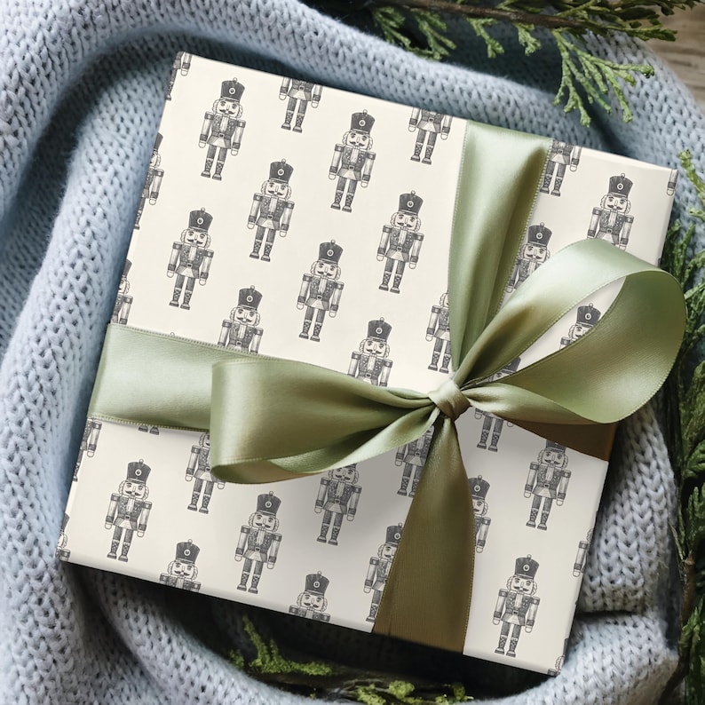 A top-down view of a present nestled in a light gray-blue knit blanket with evergreen branches placed among the blanket. The blanket, greenery, and present sit on a medium toned wood floor. The gift features a repeating pattern of traditional nutcrackers in a cream color. The nutcracker pattern is charcoal gray. The gift is wrapped in a bright sage green satin ribbon and tied into a bow.