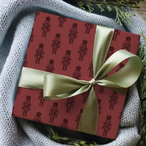 A top-down view of a present nestled in a light gray-blue knit blanket with evergreen branches placed among the blanket. The blanket, greenery, and present sit on a medium toned wood floor. The gift features a repeating pattern of etched-style nutcrackers in a burgundy color. The nutcracker pattern is a slightly darker shade of burgundy. The gift is wrapped in a bright sage green satin ribbon and tied into a bow.
