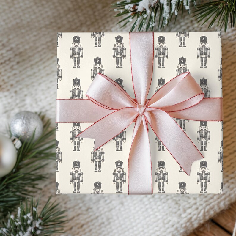 A top-down view of a gift sitting on a cream knit blanket lying on a medium toned wood floor. Flocked greenery and satin finish white and silver Christmas ornaments surround the gift. The present is wrapped in a repeating pattern of an etched-style nutcracker. The wrapping paper is cream and the nutcracker pattern is charcoal gray. The gift is wrapped in a light pink satin ribbon with a darker pink edge. The gift is tied with a bow at the center.