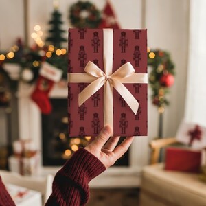 The back of a white woman’s right arm in a burgundy sweater holds up a gift in her right hand. The gift features a pattern of etched-style nutcrackers in burgundy. The nutcracker pattern is in a slightly darker shade of burgundy red. The gift has a light beige satin ribbon tied into a bow. In the background is a white fireplace covered in Christmas stockings, miniature trees, lights, and garland. Gifts are by the fireplace. The room is cream and there is tan chair with gifts on it on the right.