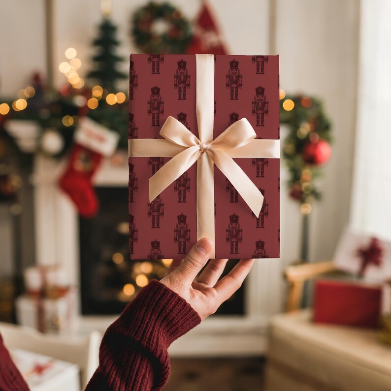 The back of a white woman’s right arm in a burgundy sweater holds up a gift in her right hand. The gift features a pattern of etched-style nutcrackers in burgundy. The nutcracker pattern is in a slightly darker shade of burgundy red. The gift has a light beige satin ribbon tied into a bow. In the background is a white fireplace covered in Christmas stockings, miniature trees, lights, and garland. Gifts are by the fireplace. The room is cream and there is tan chair with gifts on it on the right.