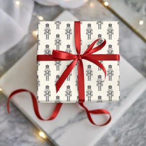 A top-down view of a wrapped gift sitting on two slabs of white and gray marble. Some sheer, white fabric and wired white fairy lights surround the gifts. The present is wrapped in a repeating pattern of an etched-style nutcracker. The wrapping paper is cream and the nutcracker pattern is charcoal gray. The gift is wrapped with a thin, red ribbon and tied with a bow.