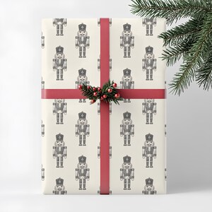 A wrapped present stands vertically on a neutral cream background. The wrapping paper features a repeating pattern of an etched-style nutcrackers in cream. The shoe pattern is a charcoal gray. The gift has a thin, red ribbon with a small sprig of greenery and red berries at the center. A branch of pine greenery is positioned in the foreground of the top right corner of the image.