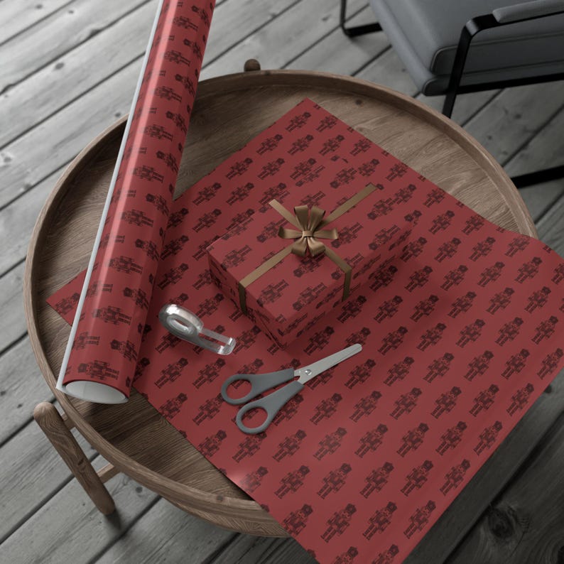 A top-down aerial view of a brown wood table sitting on a gray wood floor. On the table sits a roll and a sheet of unfurled wrapping paper in a burgundy color featuring a repeating pattern of nutcrackers. The nutcracker pattern is in a darker shade of burgundy red. On top of the sheet of wrapping paper sits a package wrapped in the gift wrap with a dark tan colored ribbon and bow. A tape dispenser and pair of scissors with a gray handle sit beside the gift to the left.