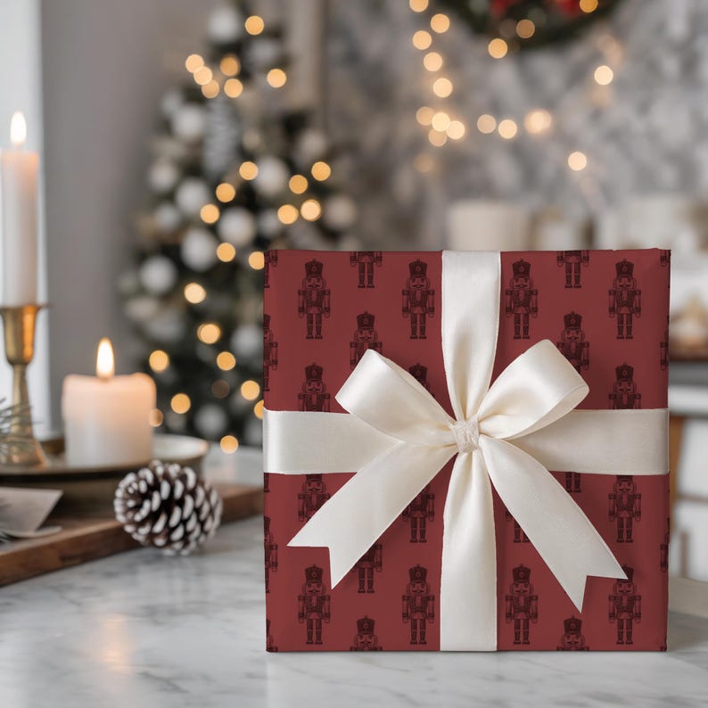 A wrapped package sits on a marble table. The wrapping paper is burgundy with a repeating pattern of etched-style nutcrackers in a darker burgundy and tied with a cream satin ribbon and bow. A wooden board with a lit white taper in a bronze candlestick, pinecones, and lit white pillar candle sits the left. The room has light gray walls and a gray wallpaper. A flocked Christmas tree with white lights sits in the corner, and a wreath are partially shown on the wallpapered wall.