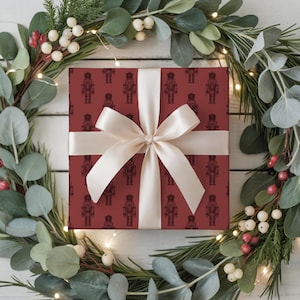 A top-down view of a wreath made of evergreen branches, eucalyptus, and red and white hypericum berries sit on a white painted wood plank floor. White fairy lights are strung around the wreath. A wrapped gift sits in the middle of the wreath. The gift is wrapped with a repeating pattern of traditional nutcrackers. The gift wrap is burgundy and the nutcracker pattern is a darker shade of burgundy. The gift is wrapped with a white satin ribbon and is tied in a bow at the center.