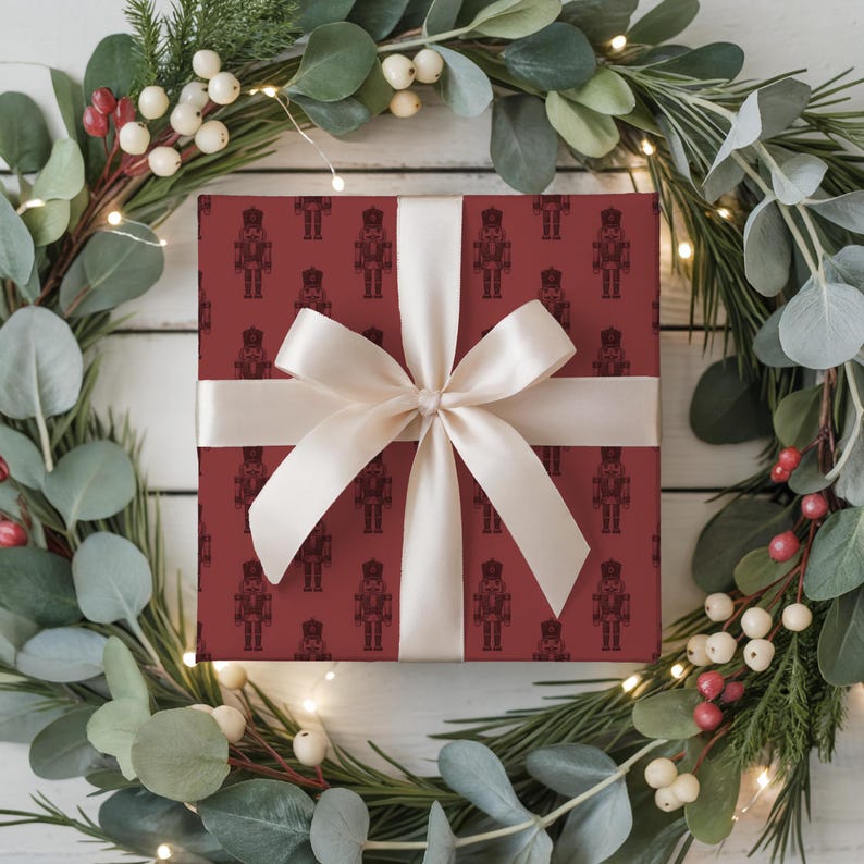 A top-down view of a wreath made of evergreen branches, eucalyptus, and red and white hypericum berries sit on a white painted wood plank floor. White fairy lights are strung around the wreath. A wrapped gift sits in the middle of the wreath. The gift is wrapped with a repeating pattern of traditional nutcrackers. The gift wrap is burgundy and the nutcracker pattern is a darker shade of burgundy. The gift is wrapped with a white satin ribbon and is tied in a bow at the center.