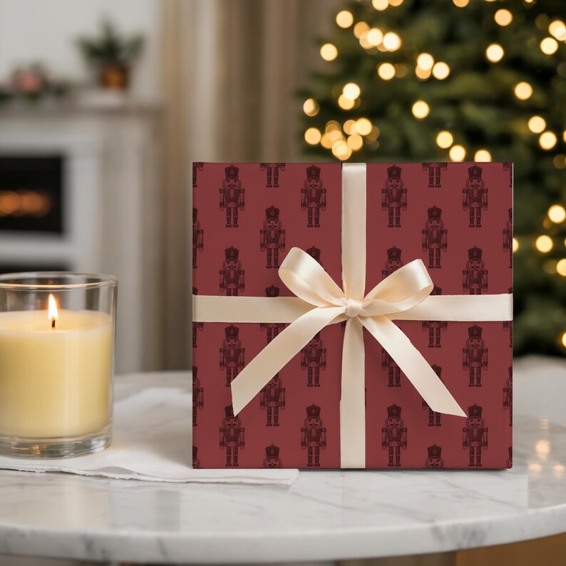 A wrapped gift sits on a marble tabletop with a cream-colored lit candle in a glass jar to the left. The wrapping paper features a repeating pattern of etched style traditional nutcrackers. The wrapping paper is burgundy, and the nutcracker pattern is in a darker shade of burgundy. The gift is wrapped with a thin, cream satin ribbon. Out of focus in the background, a white fireplace with greenery on the mantel.