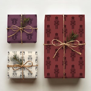 Three gifts are shown from a top-down angle on a cream background. The first box has cream wrapping paper of an etched-style pair of ballet slippers in a charcoal gray repeating pattern. The next gift features plum wrapping paper featuring etched-style images of a rat king head, wearing a crown in a darker plum. The last gift has burgundy paper with a repeating pattern of etched-style nutcrackers in a darker red. All gifts are tied with twine, and a branch of evergreen is tied into each bow.