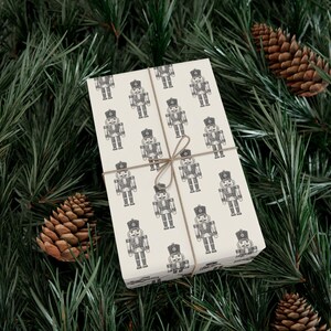 A box wrapped in cream colored wrapping paper featuring a repeating pattern of etched-style nutcrackers. The nutcracker pattern is charcoal gray, and the box is tied with tan twine. The package is lying among pine branches, and two pinecones sit to each side.