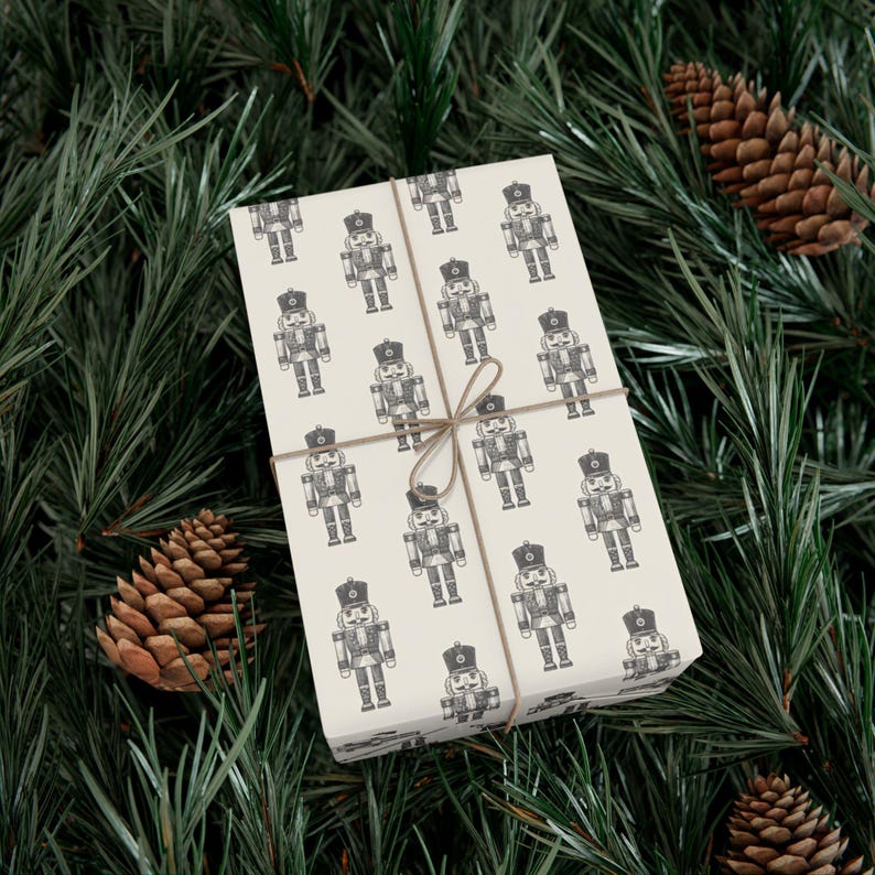 A box wrapped in cream colored wrapping paper featuring a repeating pattern of etched-style nutcrackers. The nutcracker pattern is charcoal gray, and the box is tied with tan twine. The package is lying among pine branches, and two pinecones sit to each side.