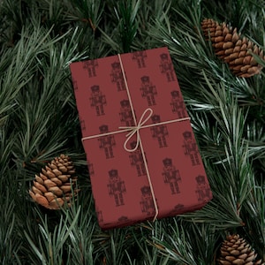 A box wrapped in burgundy colored wrapping paper featuring a repeating pattern of etched-style traditional nutcrackers. The nutcracker pattern is in a darker shade of burgundy, and the box is tied with tan twine. The package is lying among pine branches, and two pinecones sit to each side.
