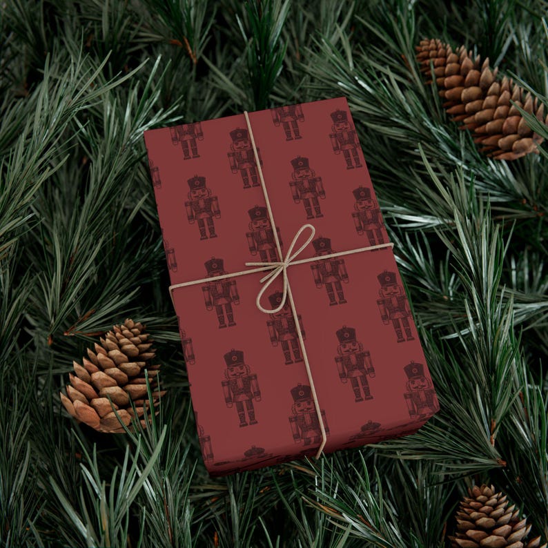 A box wrapped in burgundy colored wrapping paper featuring a repeating pattern of etched-style traditional nutcrackers. The nutcracker pattern is in a darker shade of burgundy, and the box is tied with tan twine. The package is lying among pine branches, and two pinecones sit to each side.