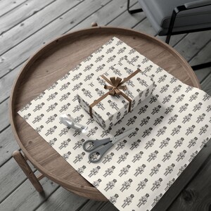 A top-down aerial view of a brown wood table sitting on a gray wood floor. On the table sits a sheet of unfurled wrapping paper in a cream color featuring a repeating pattern of traditional nutcrackers. The nutcracker pattern is in charcoal gray. On top of the sheet of wrapping paper sits a package wrapped in the gift wrap with a dark tan colored ribbon and bow. A tape dispenser and pair of scissors with a gray handle sit beside the gift to the left.