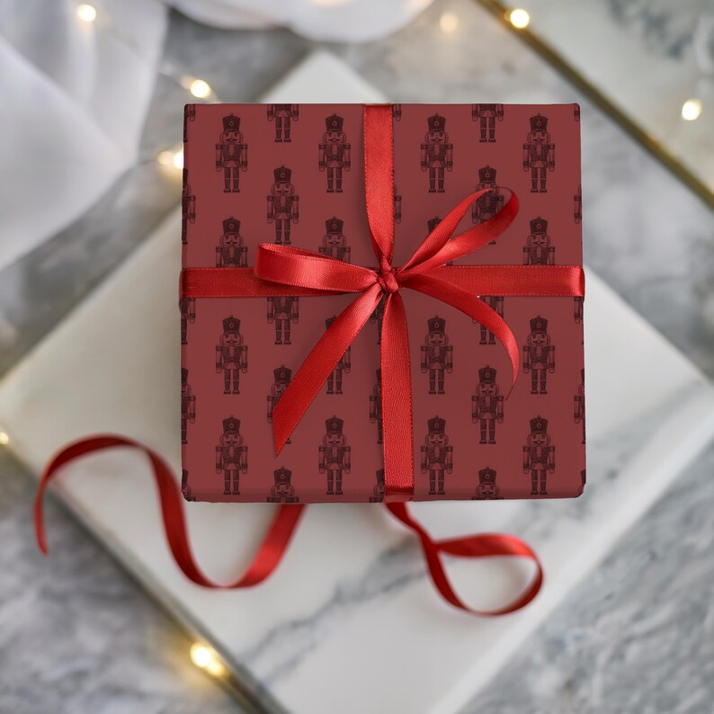 A top-down view of a wrapped gift sitting on two slabs of white and gray marble. Some sheer, white fabric and wired white fairy lights surround the gifts. The present is wrapped in a repeating pattern of an etched-style nutcracker. The wrapping paper is burgundy and the nutcracker pattern is a slightly darker red shade. The gift is wrapped with a thin, red ribbon and tied with a bow.