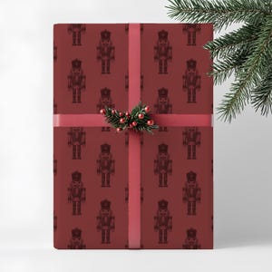 A wrapped present stands vertically on a neutral cream background. The wrapping paper features a repeating pattern of an etched-style nutcrackers in burgundy. The shoe pattern is in a darker shade of red. The gift has a thin, red ribbon with a small sprig of greenery and red berries at the center. A branch of pine greenery is positioned in the foreground of the top right corner of the image.