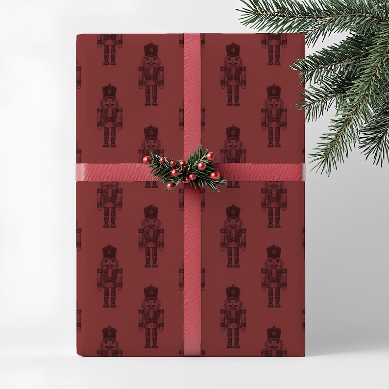 A wrapped present stands vertically on a neutral cream background. The wrapping paper features a repeating pattern of an etched-style nutcrackers in burgundy. The shoe pattern is in a darker shade of red. The gift has a thin, red ribbon with a small sprig of greenery and red berries at the center. A branch of pine greenery is positioned in the foreground of the top right corner of the image.