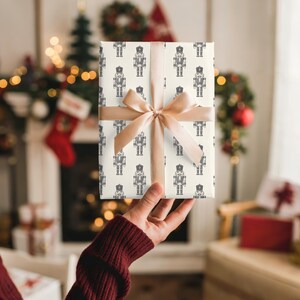 The back of a white woman’s right arm in a burgundy sweater holds up a small gift in her right hand. The gift features a repeating pattern of an etched-style nutcrackers in cream. The nutcracker pattern is in charcoal gray. The gift has a light beige satin ribbon tied into a bow.  In the background is a white fireplace covered in Christmas stockings, miniature trees, lights, and garland. Gifts are by the fireplace. The room is cream and there is tan chair with gifts on it on the right.