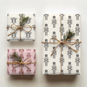 Three gifts are shown from a top-down angle on a cream background. The first box features pink wrapping paper of an etched-style pair of ballet slippers in a darker pink repeating pattern. The next gift features cream wrapping paper featuring etched-style images of a rat king head, wearing a crown in charcoal gray. The last gift has cream paper with a repeating pattern of etched-style nutcrackers in charcoal gray. All gifts are tied with twine, and a branch of evergreen is tied into each bow.