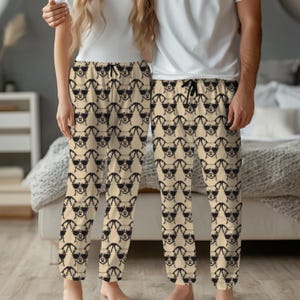 May include: Tan pajama trousers featuring a repeating pattern of black dog faces wearing sunglasses. The trousers have a drawstring waist. The background includes a bed and a nightstand.