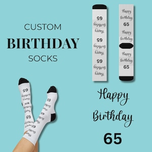 Pode incluir: Meias de aniversário brancas personalizadas com detalhes em preto e o texto "Happy Birthday 65". As palavras "Custom Birthday Socks" são exibidas em preto. As meias estão em um fundo azul claro.