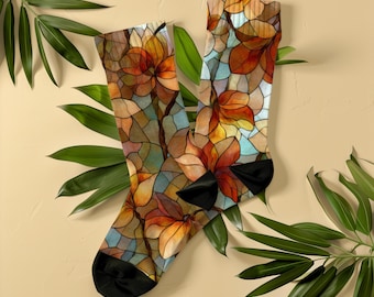 Könnte beinhalten: Ein Paar Socken mit einem floralen Buntglasdesign. Die Socken zeigen ein Muster aus orangefarbenen, braunen und gelben Blumen mit blauen Akzenten. Die Zehen und Fersen sind schwarz. Die Socken werden auf einem beigefarbenen Hintergrund mit grünen Blättern präsentiert.
