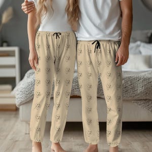 May include: Beige pajama pants with a repeating black line drawing of a dog's face. The pants have a drawstring waist. The person on the left is wearing a white top, and the person on the right is wearing a white top.
