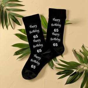 Pode incluir: Um par de meias pretas com a frase "Happy Birthday 65" em branco. As meias são exibidas em uma superfície bege clara com folhas verdes.