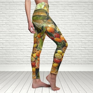 Op de afbeelding: Leggings met een levendig fruitpatroon met sinaasappels, citroenen en andere citrusvruchten. De leggings hebben een hoge taille en een kleurrijk, mozaïekachtig ontwerp. De achtergrond is een witte bakstenen muur en een lichte houten vloer.