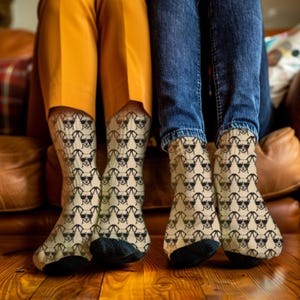 May include: A pair of socks with a repeating pattern of a dog wearing sunglasses. The socks are beige with black and white dog graphics. The socks are worn by two people, one wearing yellow pants and the other wearing blue jeans.