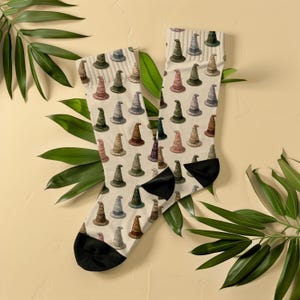 Könnte beinhalten: Ein Paar Socken mit einem durchgehenden Muster aus kleinen, spitzen Zauberhüten in verschiedenen gedeckten Farben wie Grün, Rosa, Braun und Grau auf cremefarbenem Grund, ergänzt durch abgesetzte schwarze Zehen- und Fersenbereiche.