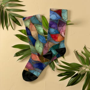 May include: A pair of colorful socks with a stained glass design. The socks feature a pattern of blue, green, orange, and purple shapes outlined in gold. The toes and heels are black. The socks are displayed on a beige background with green leaves.