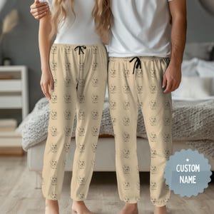 Könnte beinhalten: Beige Pyjamahose mit schwarzem Kordelzug und einem sich wiederholenden Muster von Hundeillustrationen. Die Hose ist Teil eines passenden Sets, ideal zum Faulenzen oder Schlafen. Die Pyjamahose ist aus einem weichen, bequemen Material.