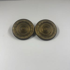 Può includere: Due pomelli per mobili antichi color ottone. Ogni pomello è rotondo con un design circolare centrale e anelli concentrici. Il bordo esterno presenta un motivo a volute decorativo. I pomelli sono esposti su una superficie bianca.