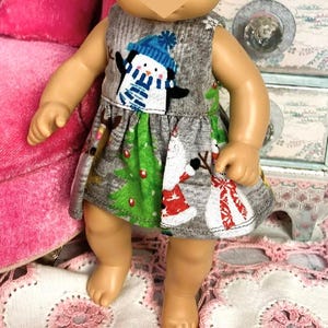 May include: A doll wearing a gray dress with a festive penguin and Christmas tree design. The doll also has a matching bow headband. The dress features a green skirt with a snowman and Christmas tree print. The doll is standing on a pink and white floral doily.